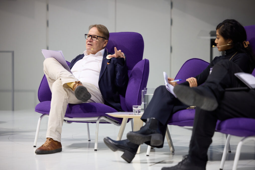 Harri Ohra-aho seated on a purple seat on a stage beside another speaker.